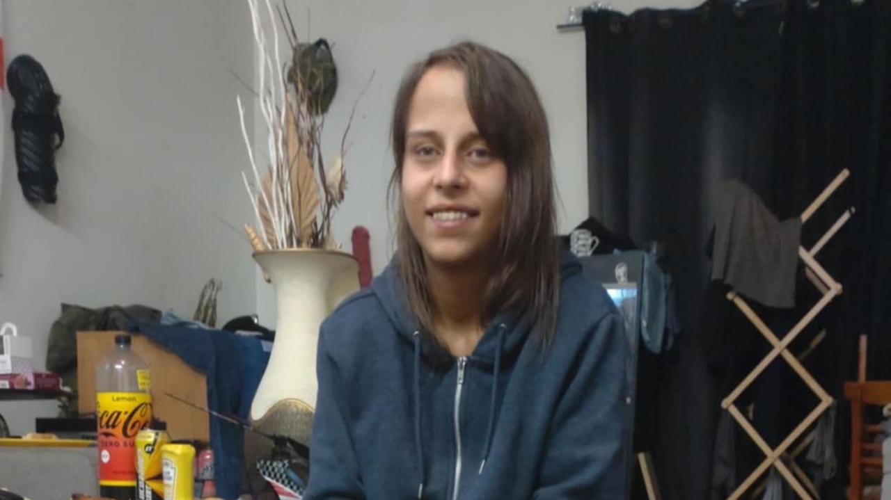 A woman with long brown hair wearing a blue hooded top with a zip. She is smiling but not looking into the camera. There is a clothes horse, a vase and various cans and plastic bottles in the background.