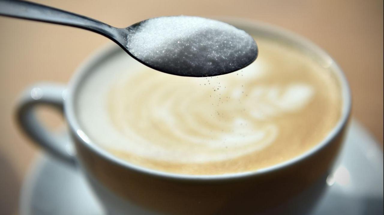 A teaspoon of white sugar being held over a cup of milky coffee with grains of sugar falling into the cup. The cup and saucer are both white.