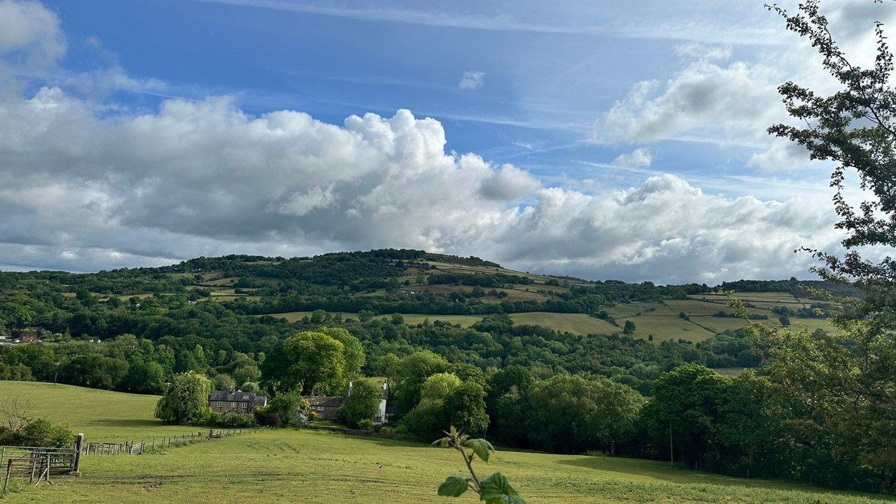 A green field with houses in distance, bushy trees and rolling countryside under a blue, cloudy sky