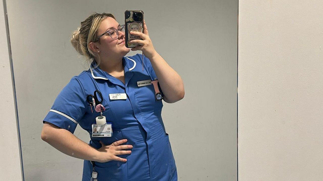 Holly is in her blue nurses' uniform and is proudly taking a selfie with her iPhone.