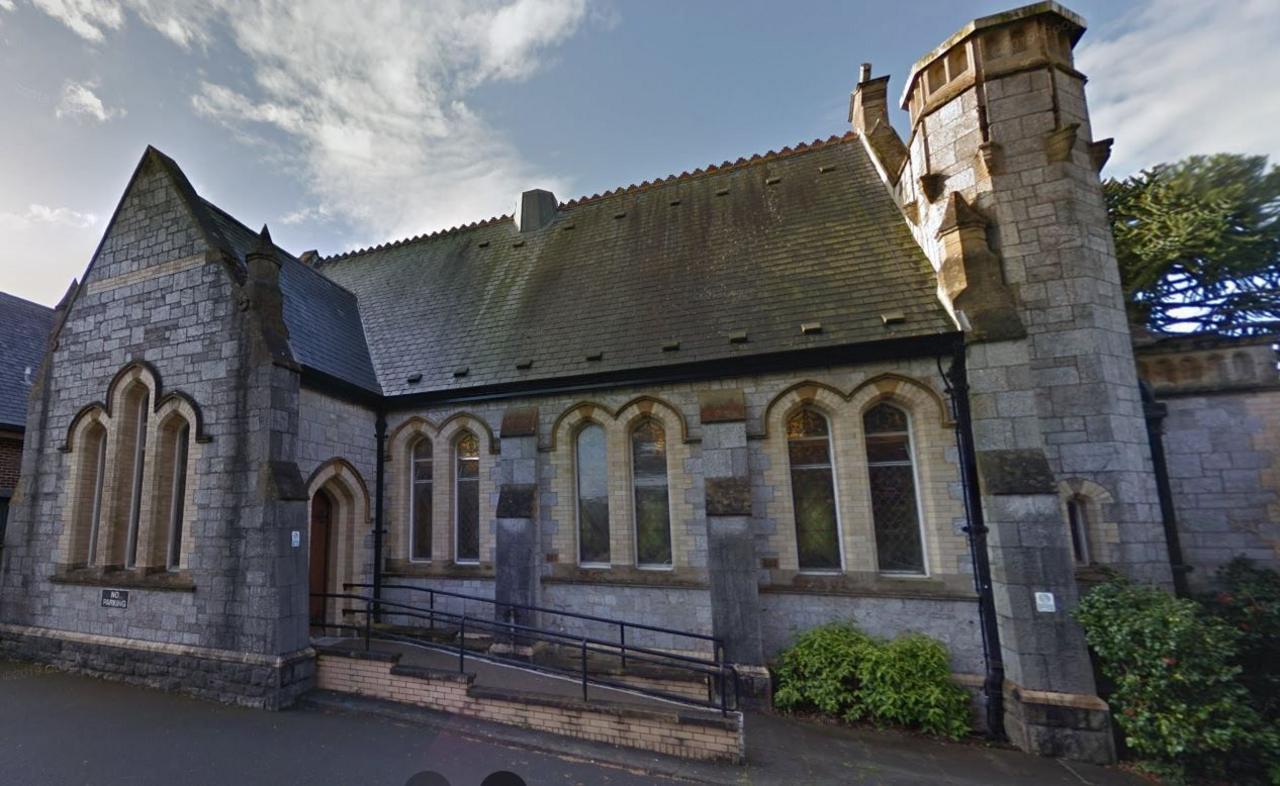 The crematorium building at Weston Mill in Plymouth. It is a stone building with Gothic architectural features, including pointed arch windows and doorways, with grey stone walls and a sloping roof covered in dark shingles. A turret-like tower is on the right side, while the windows are tall and narrow, with decorative arches at the top. It is under a partly cloudy sky.