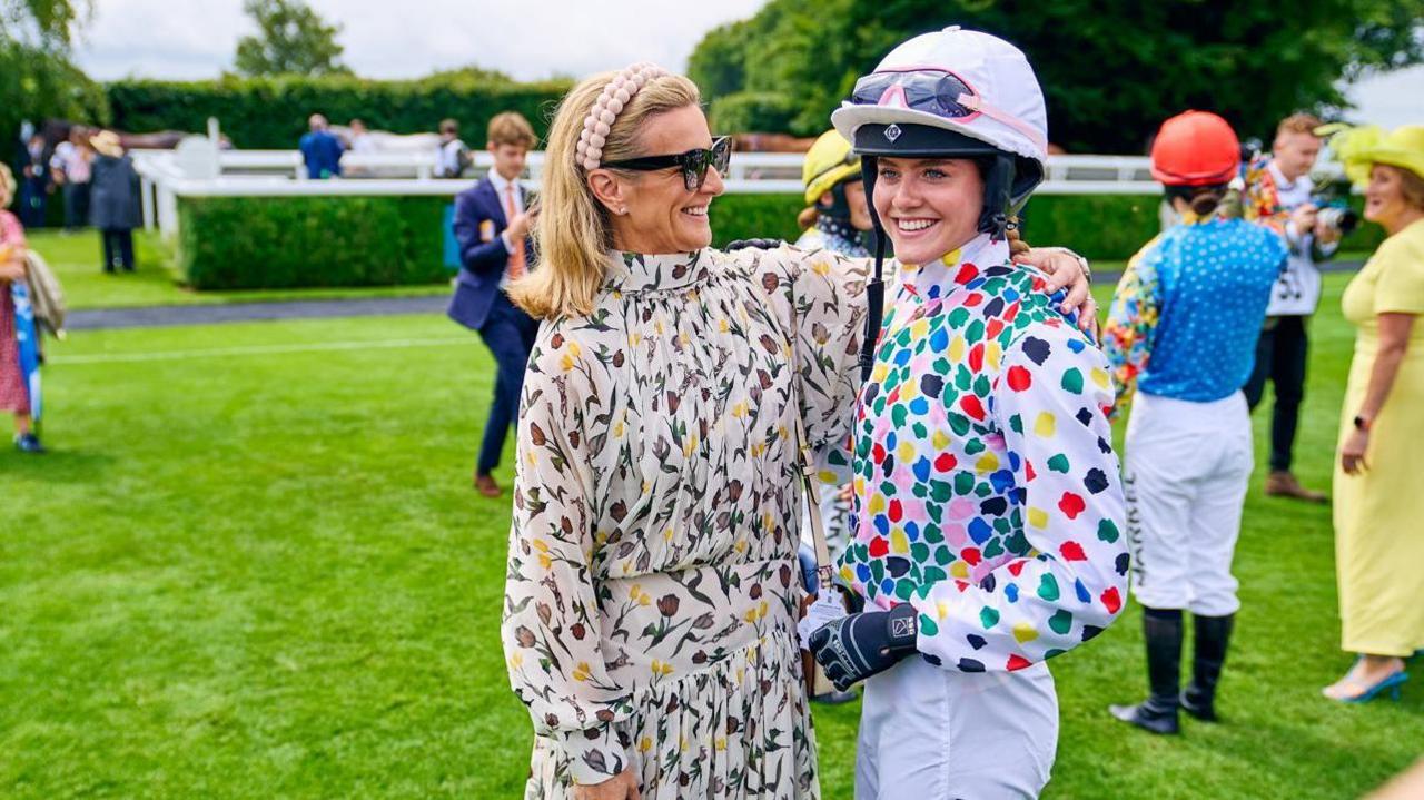Two women stood hugging. One is wearing a dress and looking at the other, while the other is looking slightly towards the camera and wearing a jockey outfit. They are stood on grass.