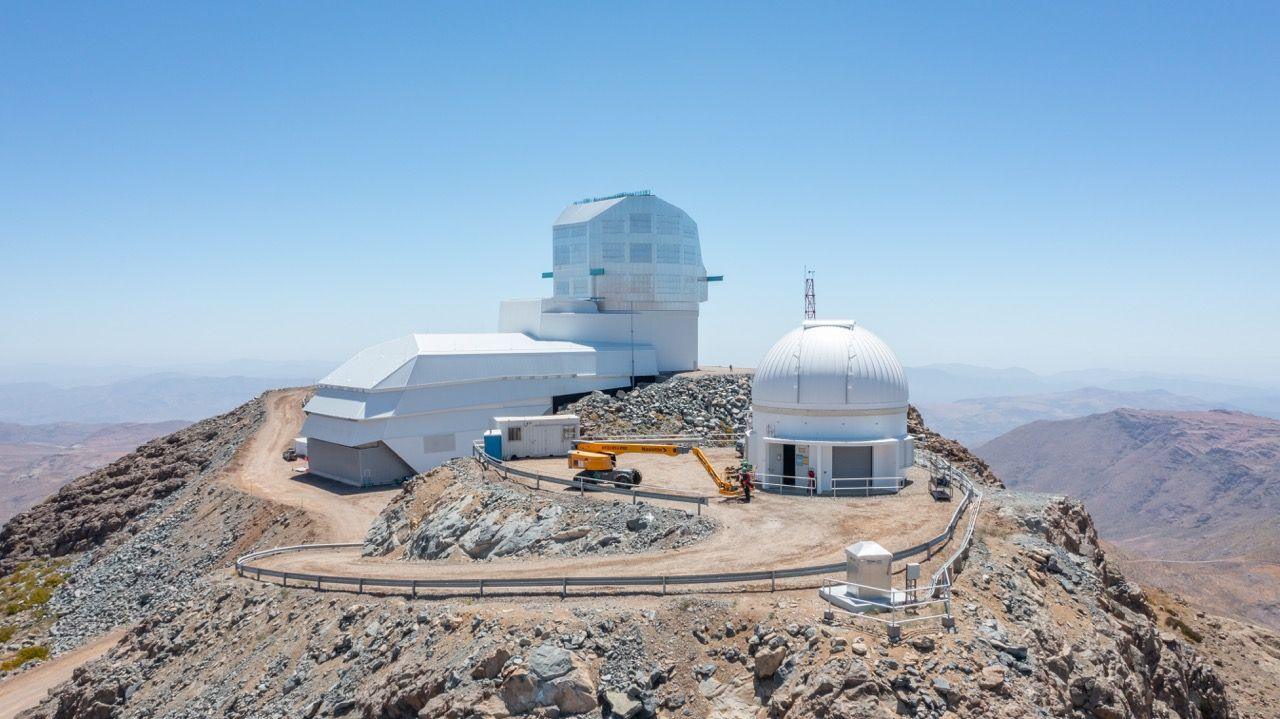 Three large white buildings stand on top of a dry mountain in a desert. One has a domed roof. In the background the sky is blue and looks dry. A yellow crane is in front of the three white buildings which are the Vera Rubin observatory. A dusty road leads up to the buildings.