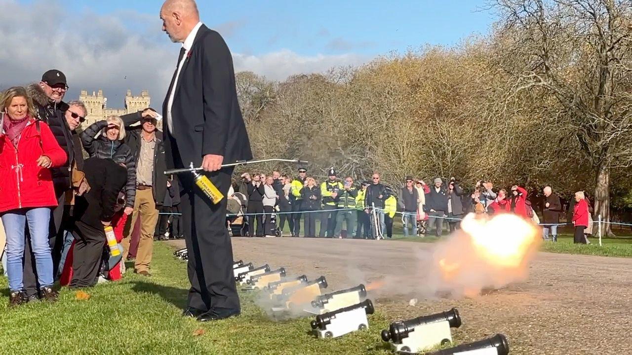 A small cannon, about the size of a brick, at the point of explosion, with an orange fireball about two feet away. There's a whole row of the small cannons, with crowds of people watching from behind a cordon. A man in a suit stands next to the cannon that has exploded.