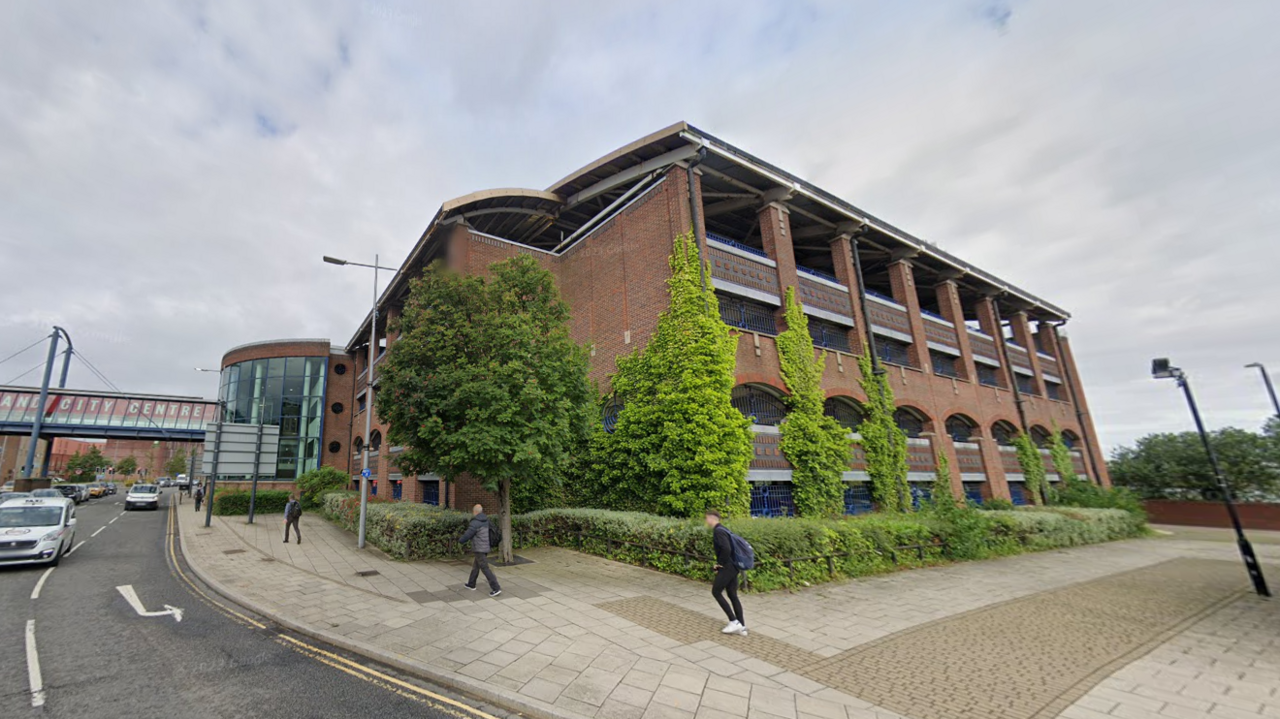 The four-storey St Mary's multi-story car park clad in red bricks with multiple large gaps in the wall. The sky is grey. A road runs to the left of it with cars coming towards the camera. A few people are walking on the street. In the distance is a covered bridge over the road with the worlds Sunderland City Centre written out across its windows.
