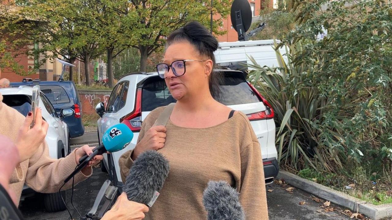 A woman in a brown jumper with brown hair tied up in a bun, speaking to reporters who have microphones and mobile phones near her. There are cars in the background and a court building.