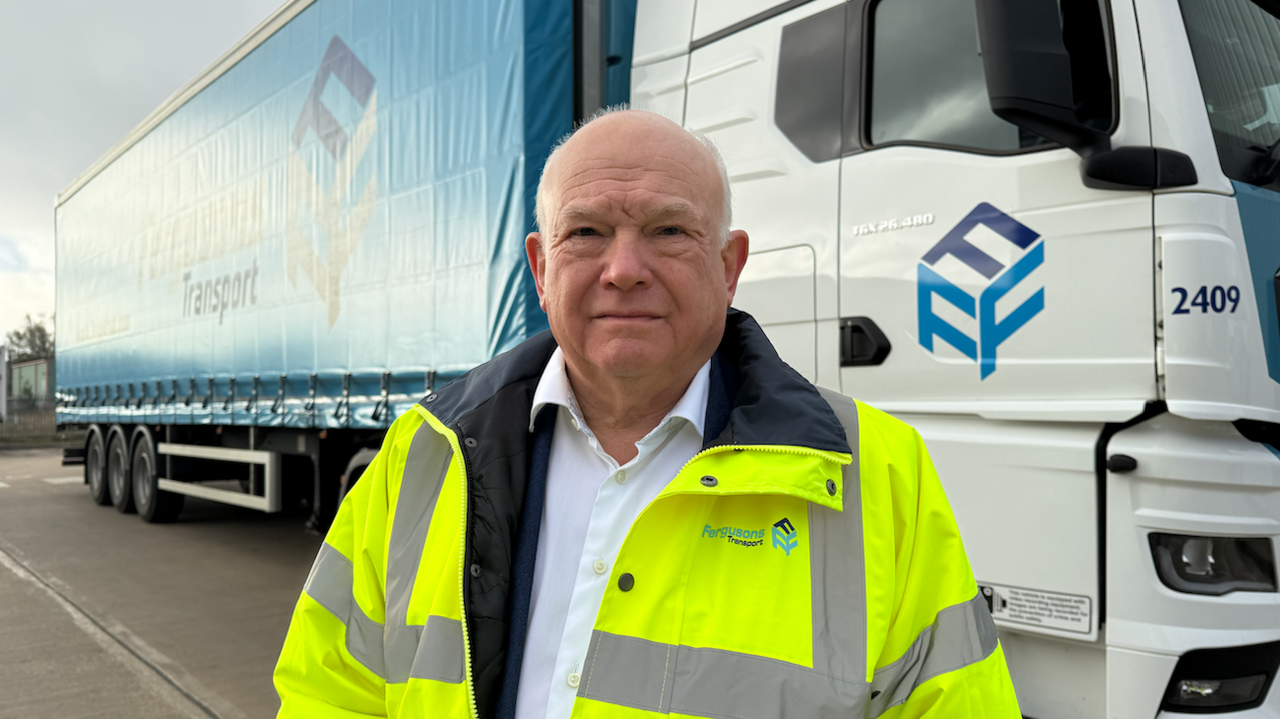 A man wearing a white shirt covered by a yellow high visibility jacket stands in front of a white and blue lorry which has Fergusons Transport written on the side.