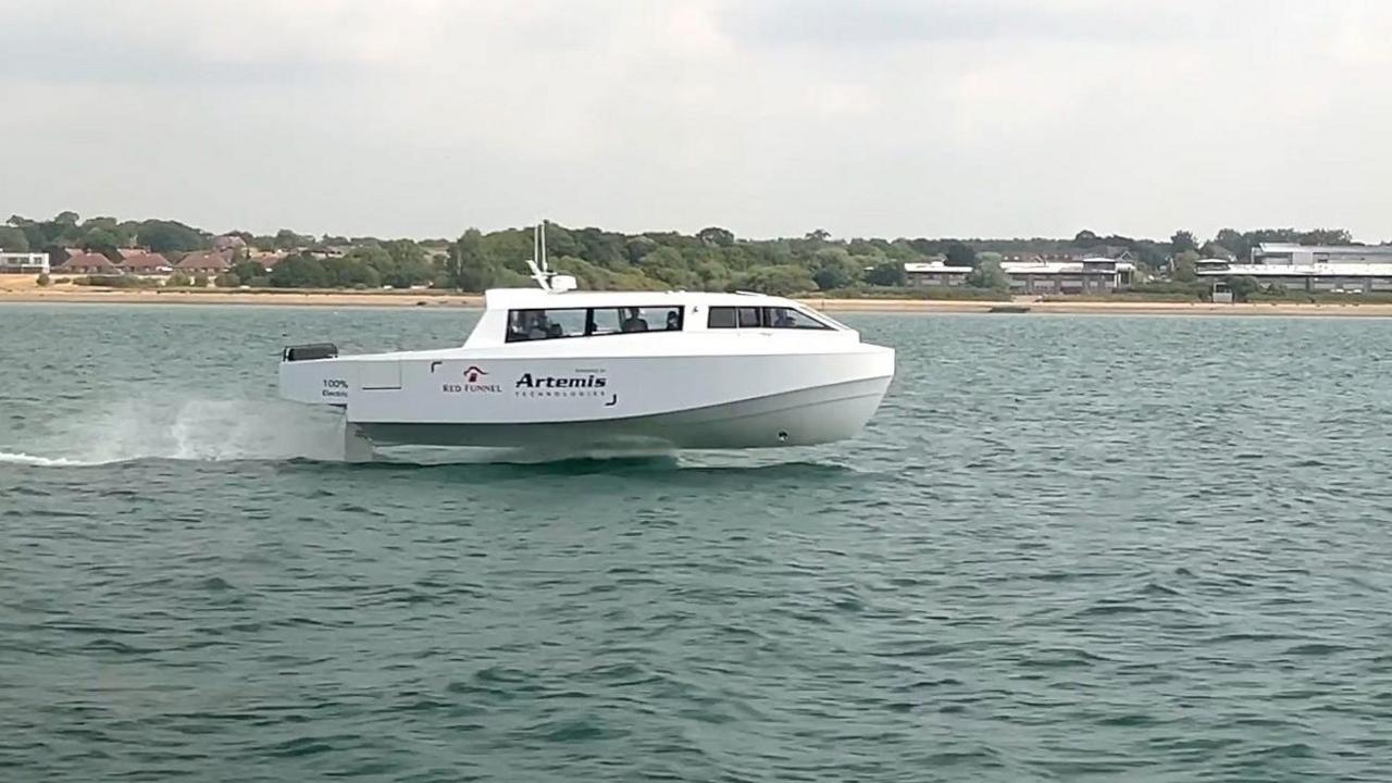 A small, sleek white vessel traveling across the water. Its hull is lifted out of the water and it appears to be gliding. In the background is the shore, with sand giving way to trees and buildings