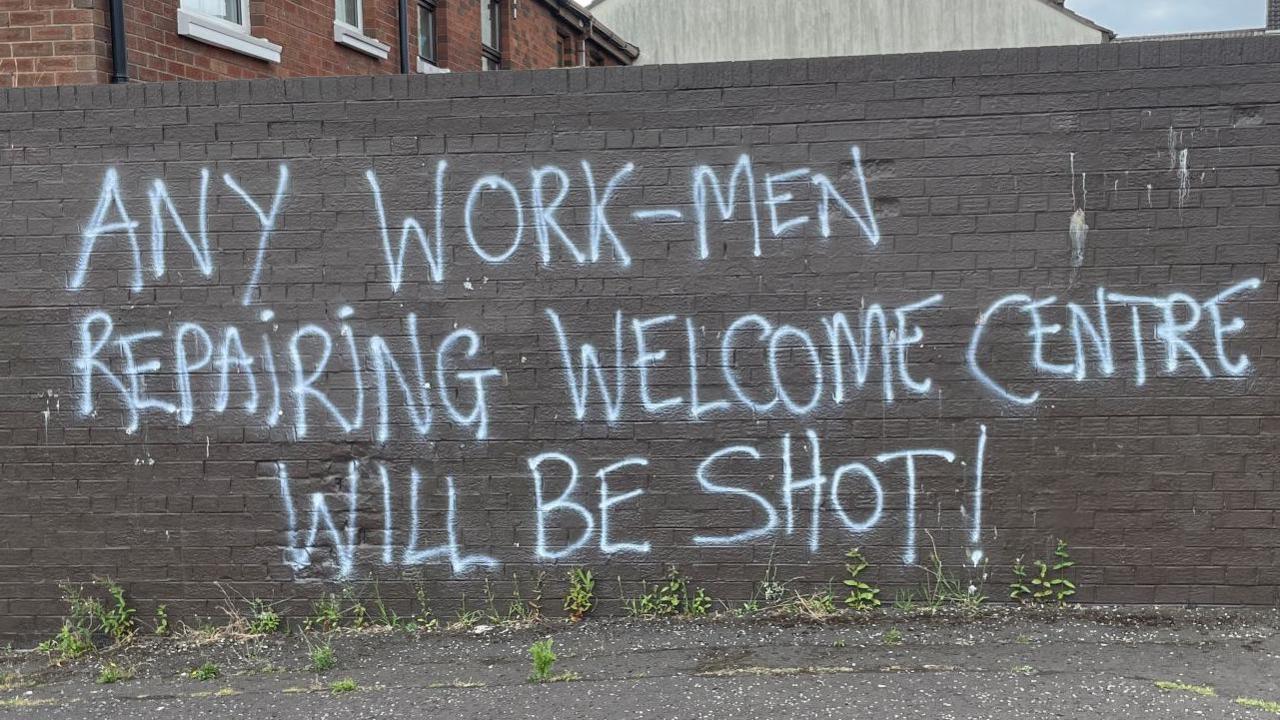 A brown mural with graffiti saying 'Any work-men repairing Welcome Centre will be shot'