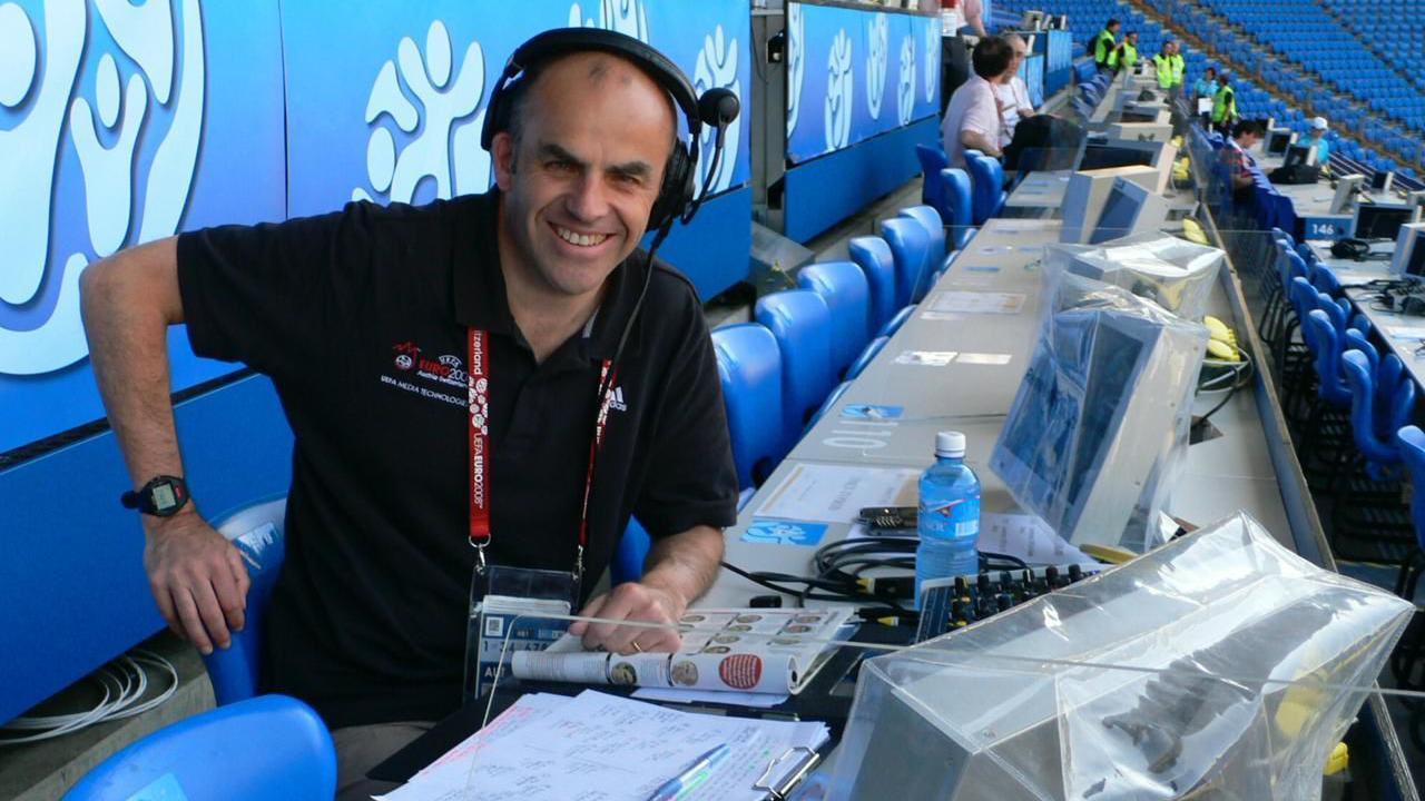 Tony Jones wearing a black polo shirt and headset while sitting in the press box at a Euro 2008 match. He is smiling and surrounded by blue seats, with TV monitors and notepads in front of him.