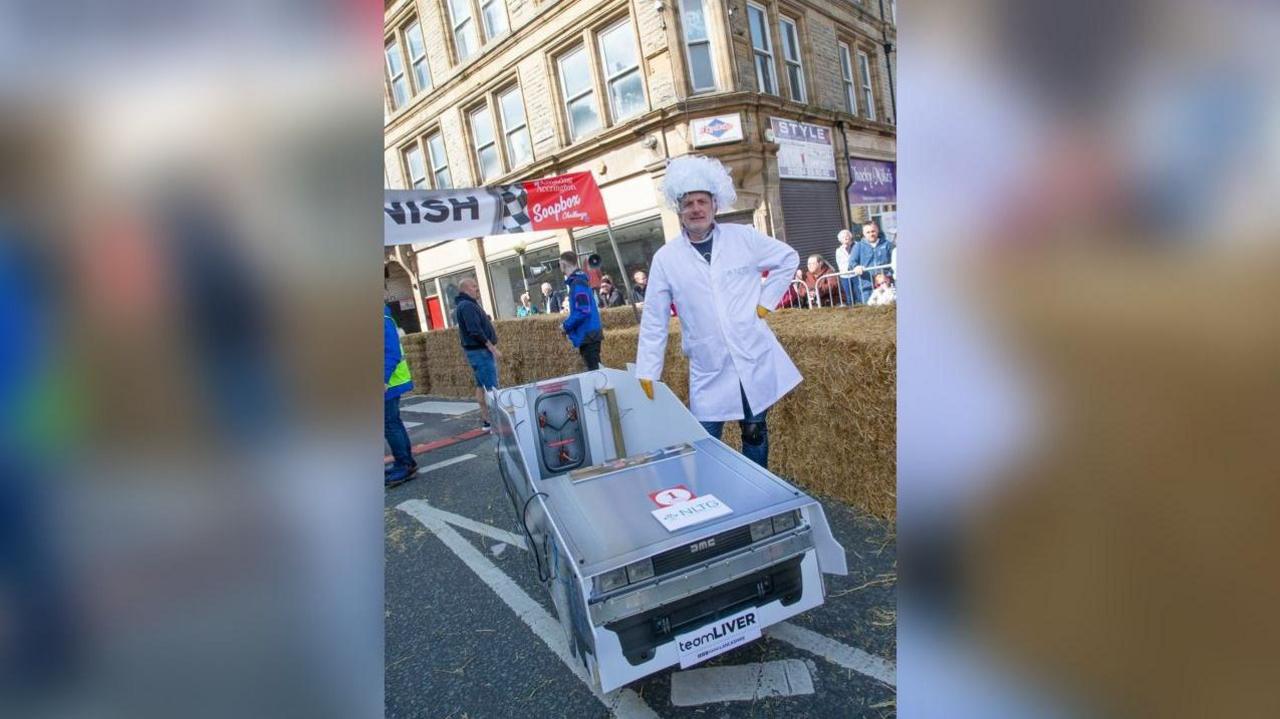 Graham Liver wears a curly white wig and white scientist-style jacket, standing next to the Delorean / Back to the Future-themed cart.