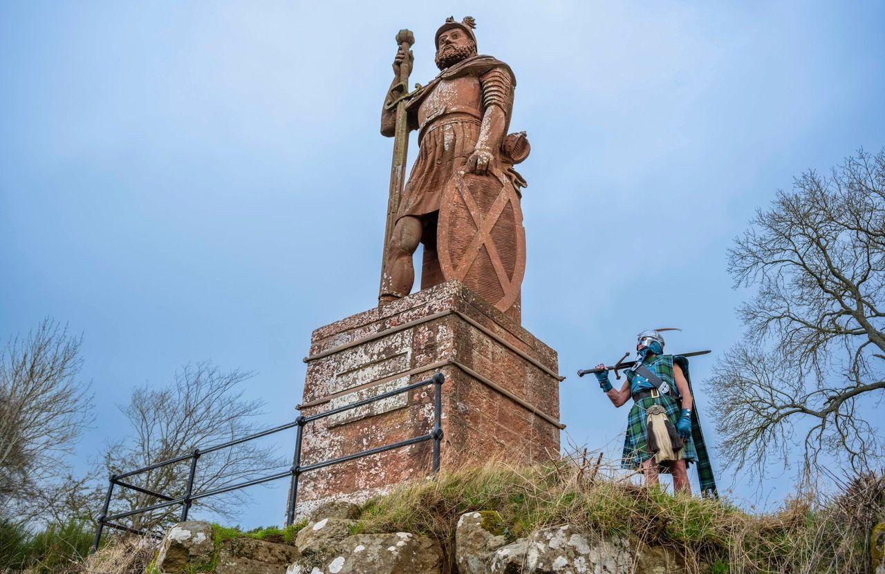 Jesse Rae in tartan dress with a claymore and helmet is dwarfed by a large sandstone statue of William Wallace