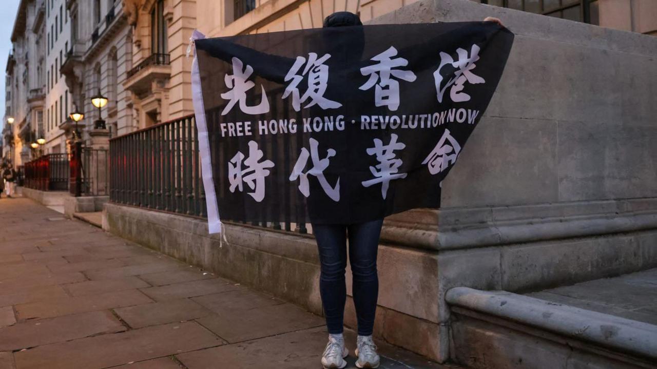 A woman holding a protest banner up made from black cloth which reads Free Hong Kong. Revolution Now. The sign also has Chinese characters on it painted in white. The banner is covering the woman's face, but  her legs in jeans and white trainers can be seen. She is in a London street, lined with multi-storey grey stone buildings with black railings at street level.