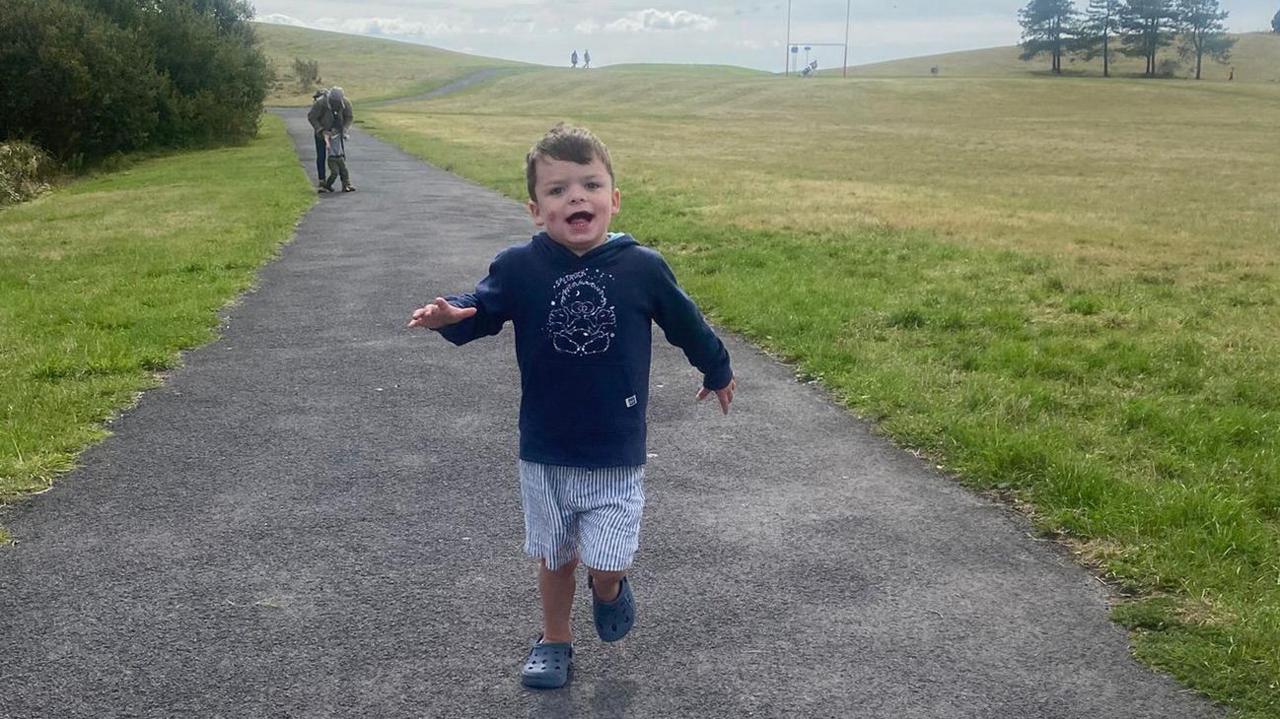 A young boy is smiling while running along a path in the countryside. He is wearing a blue hooded top and blue and white striped shorts and blue croc shoes. There are shrubs and trees in the distance. 