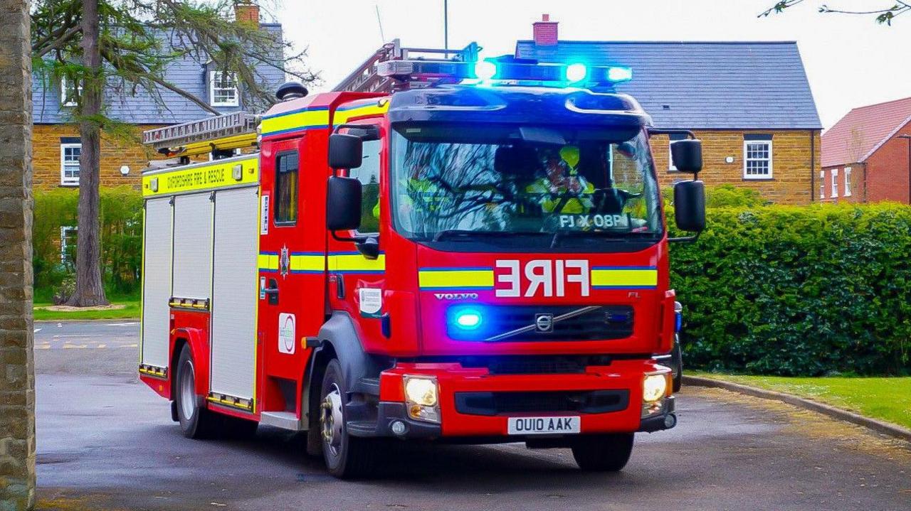 A red fire engine with its blue lights flashing travelling on a road, with new-build houses behind it.