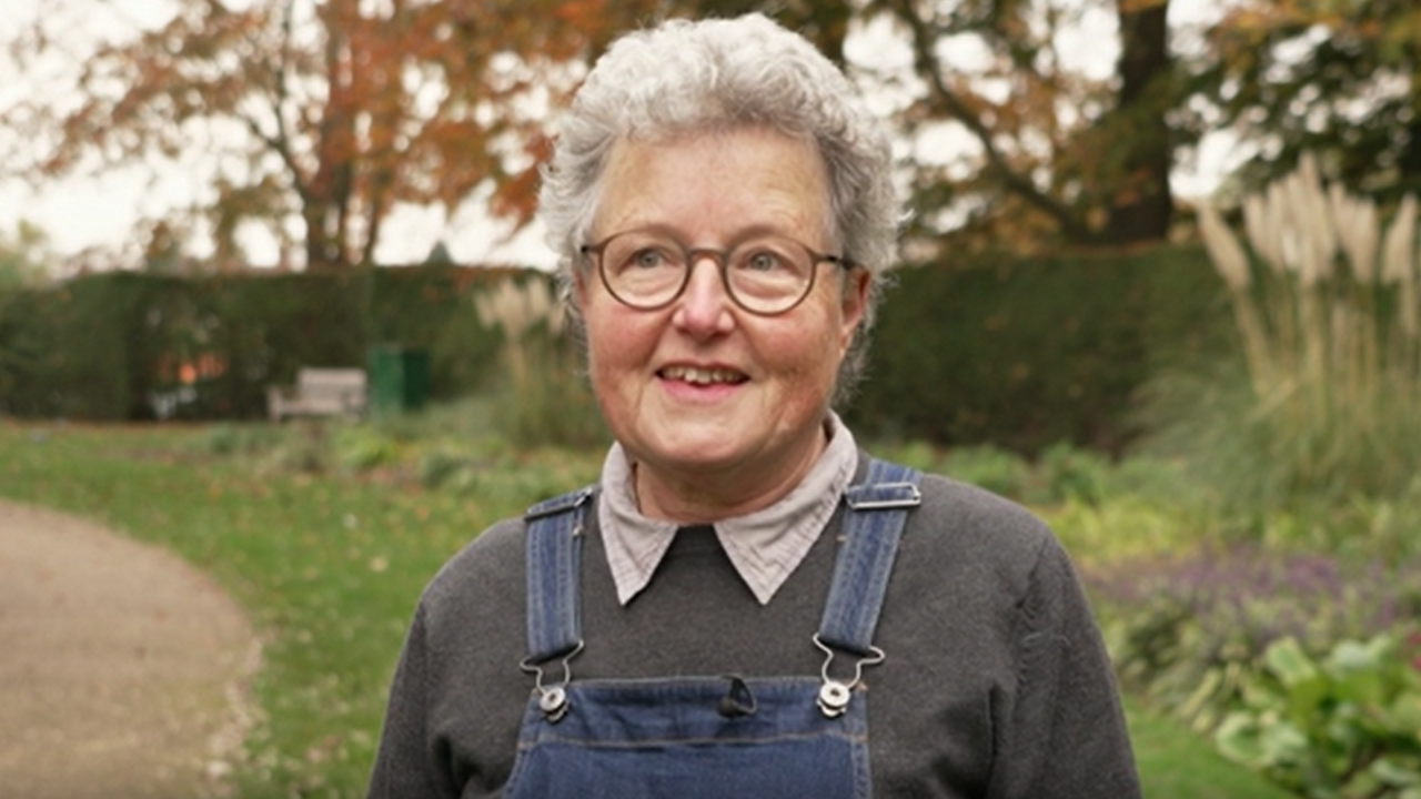 Catherine Rowett in a grey jumper and denim overalls. She is looking at the camera and has grey hair and is wearing brown rimmed circular glasses.