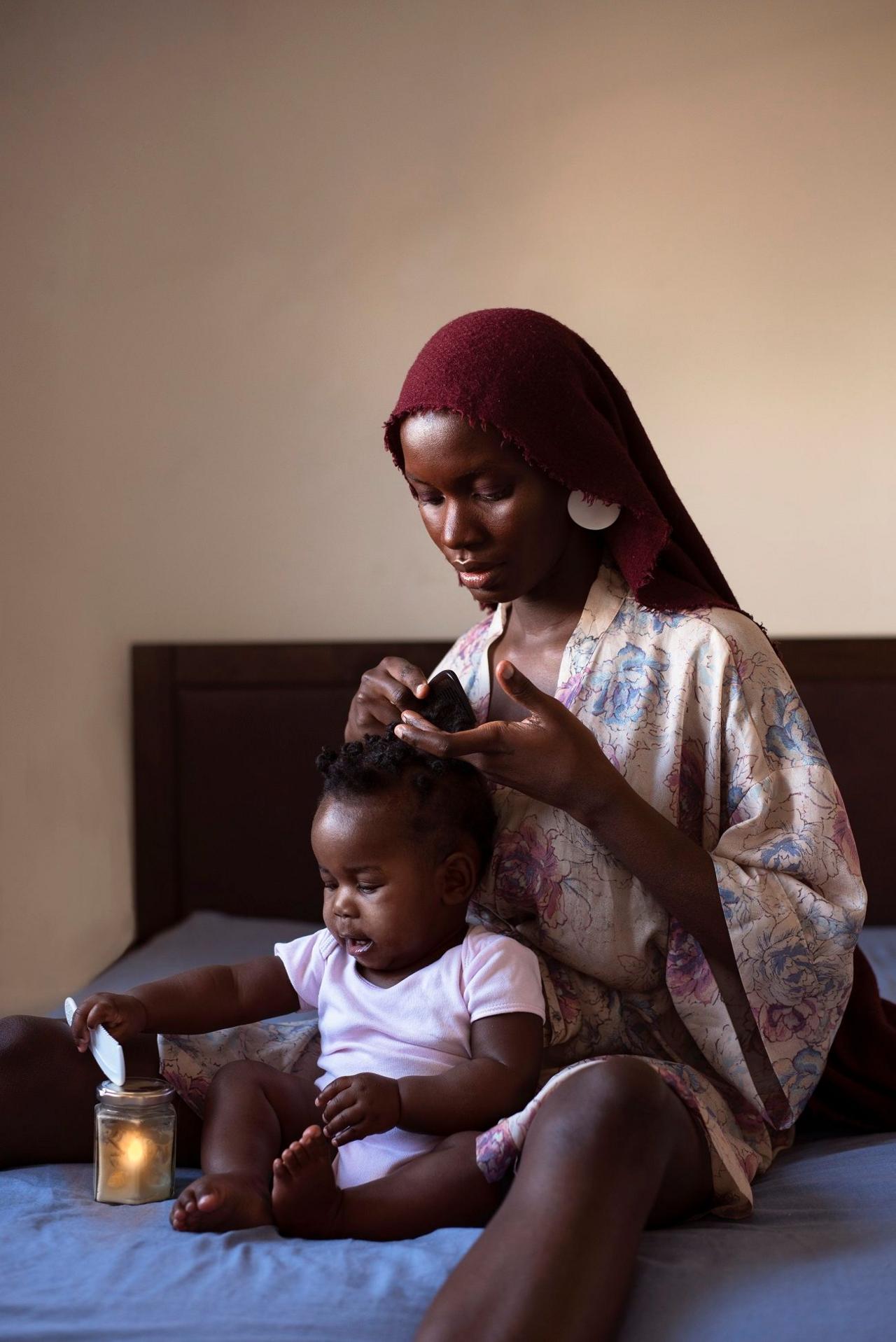 Dola Posh, with a red head covering, combs her daughter's hair as they sit on a bed with a blue covering. There is a candle in a jar next to them.