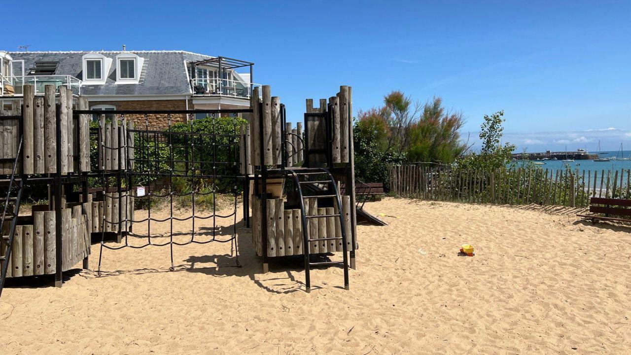 A park on sand. There is a wooden climbing frame. Behind the park is a house. The sky is blue. In the background to the right is the sea and sailing boats.