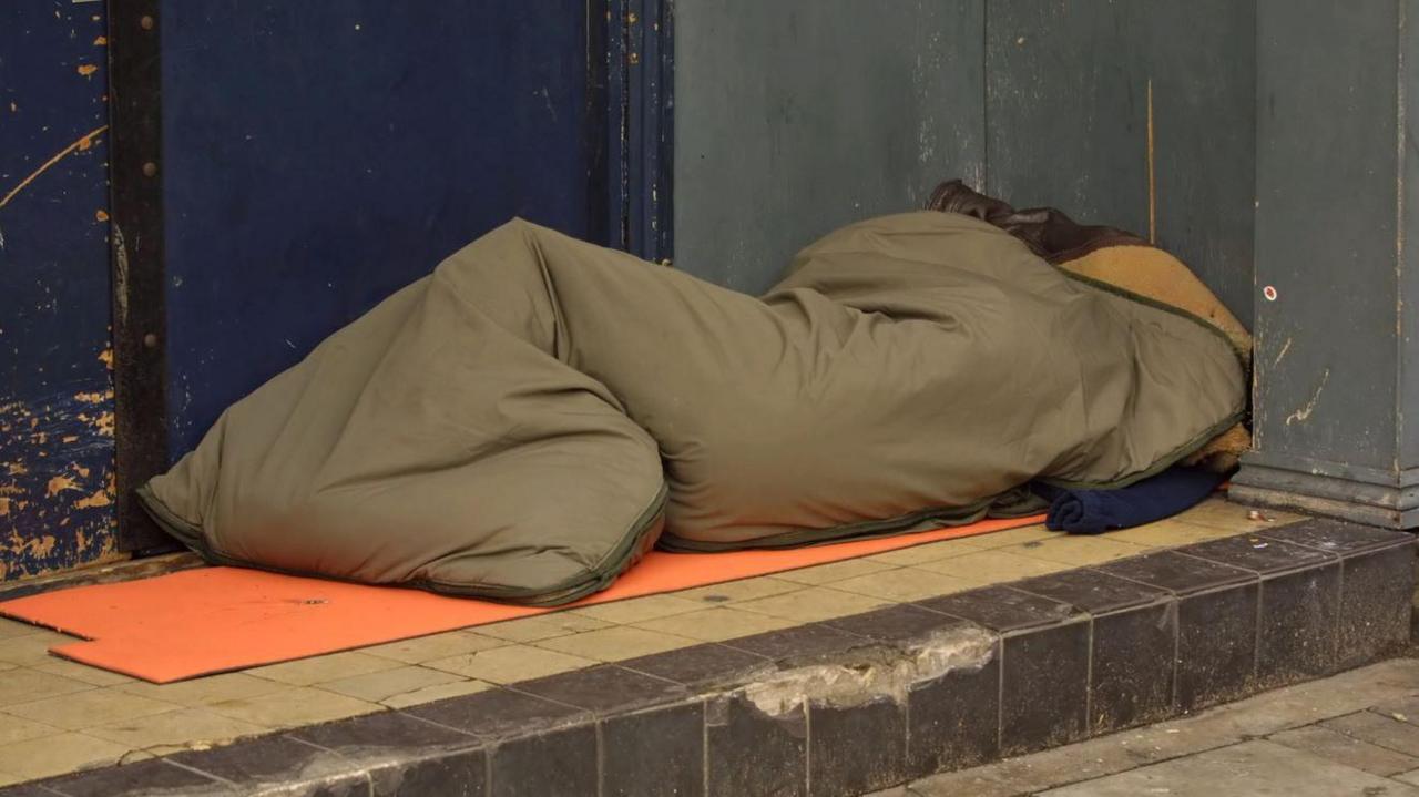 A khaki sleeping bag on a step outside a building with a blue door with someone appearing to be in it. An orange cardboard sheet is underneath the sleeping bag