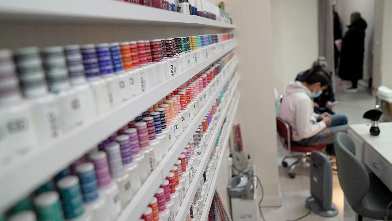 Rows of nail varnish products are seen in a shop with an anonymous nail technician working in the background