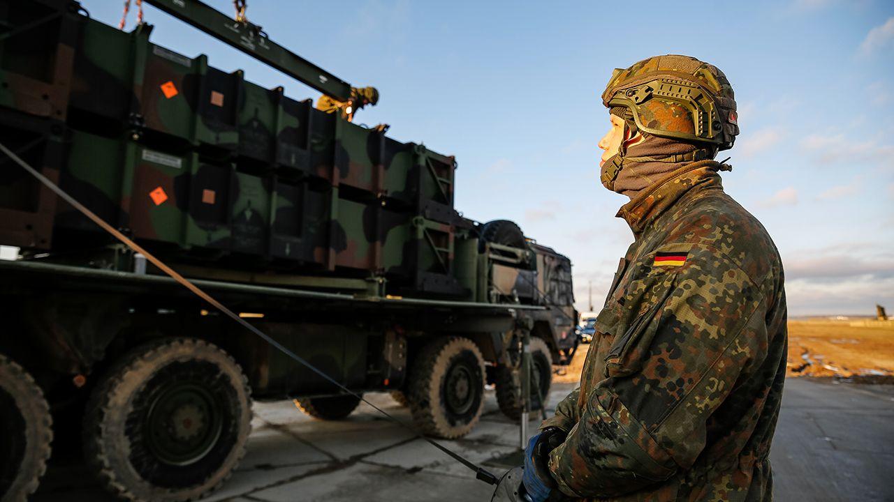 A soldier with a German flag patch sewn into the arm of their uniform operates a remote while looking up at a large military truck moving along a road against a blue sky, as German military moved air defence units to southeast Poland in January.