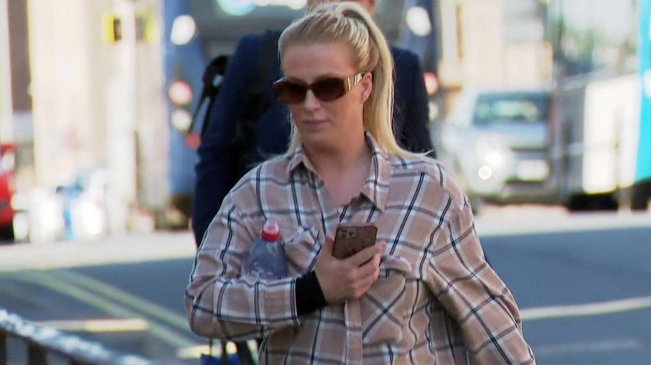 A woman with long blonde hair tied up in a ponytail, with a beige checked shirt and sunglasses, arrives at court. She has a water bottle tucked under her arm and is holding an iPhone.