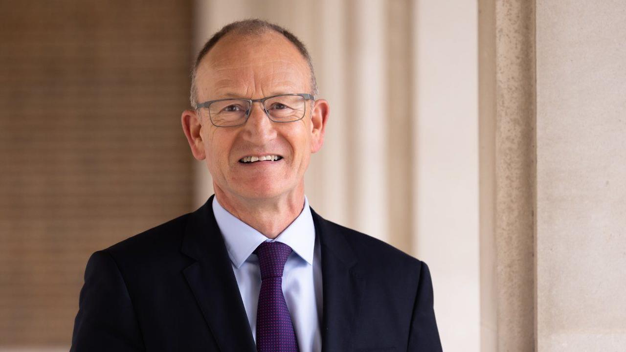 A picture of Hertfordshire County Council leader Richard Roberts wearing a black suit jacket, a purple tie, and glasses.