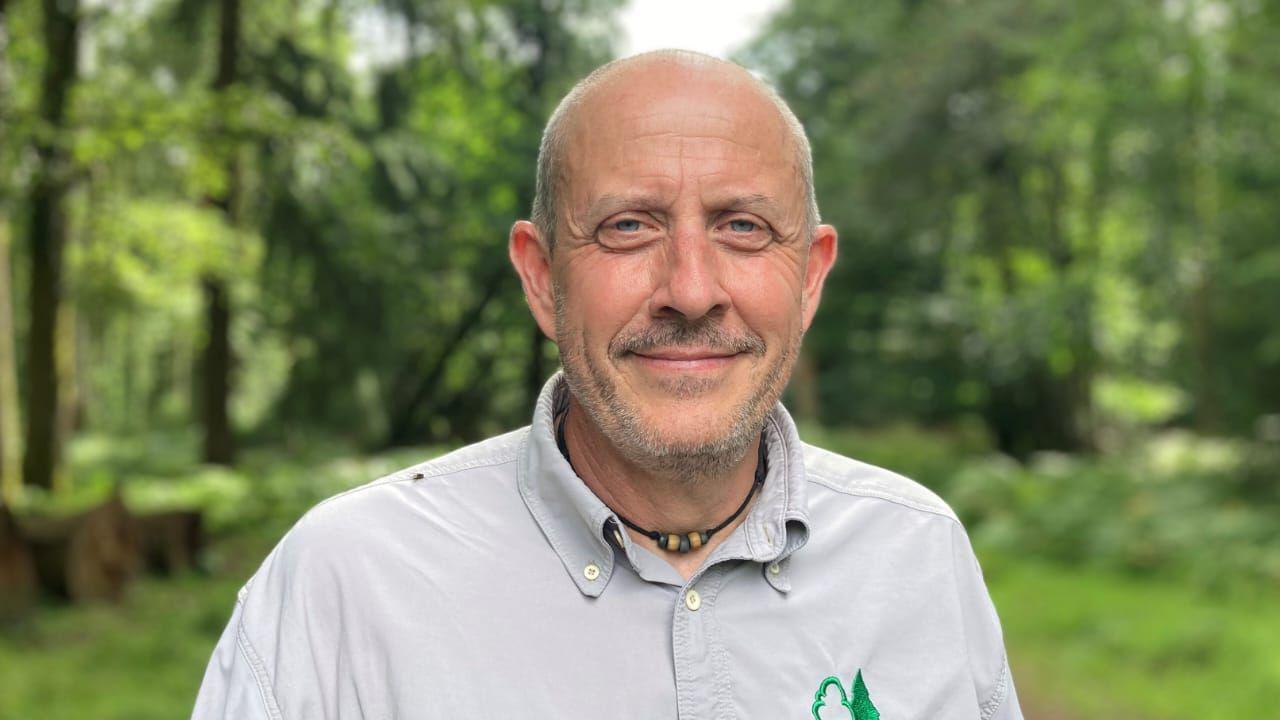 Gareth Hopkins in a forest. The background is blurred. He has a shaved head and is wearing a grey top. 