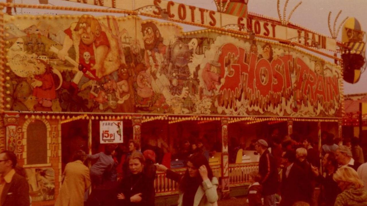 A picture with a red filter from the 1970s shows a ghost train ride with people standing outside, ready to get on.