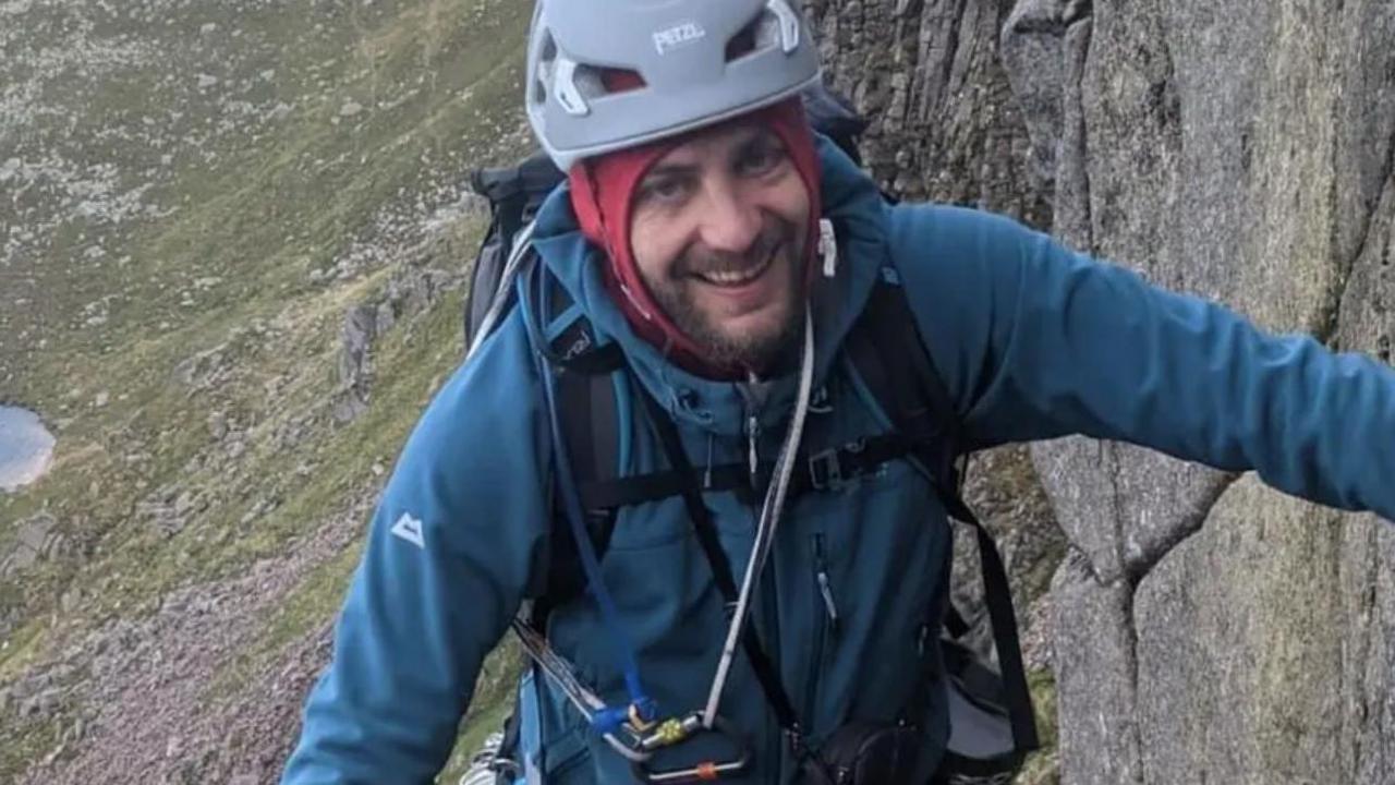 A picture of a bearded man in climbing gear, positioned next to a wall of rock. He wears a grey helmet and blue sweater. He smiles at the camera.