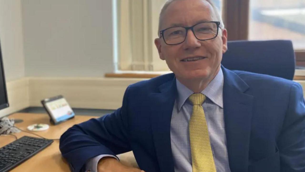 A man with short hair and a blue jacket with a light blue shirt and yellow tie sat with one arm on a desk with a computer keyboard on it