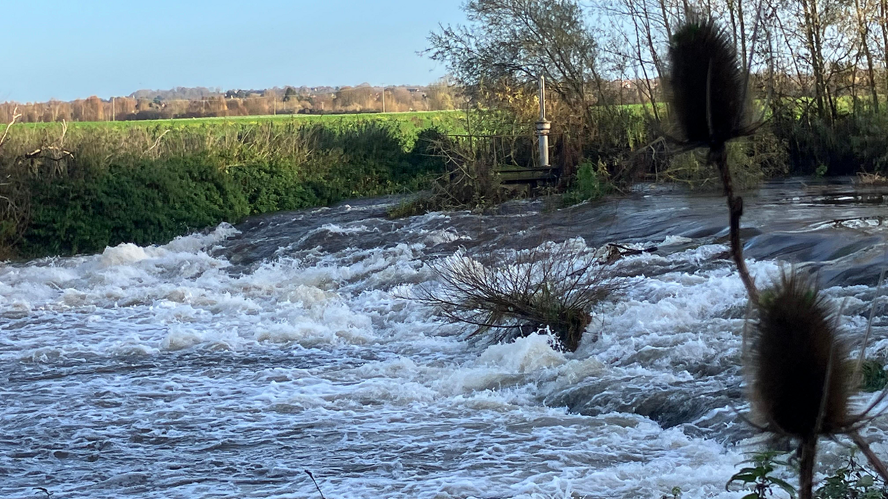 Water rushing over a weir in Bidford-on-Avon. There are green fields beyond the far bank
