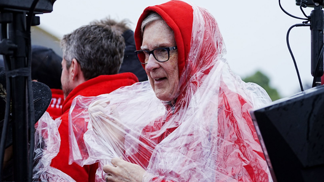 Liverpool owner John Henry in a red hoody. He is wearing glasses. He has a piece of plastic over him to protect him from any rain.