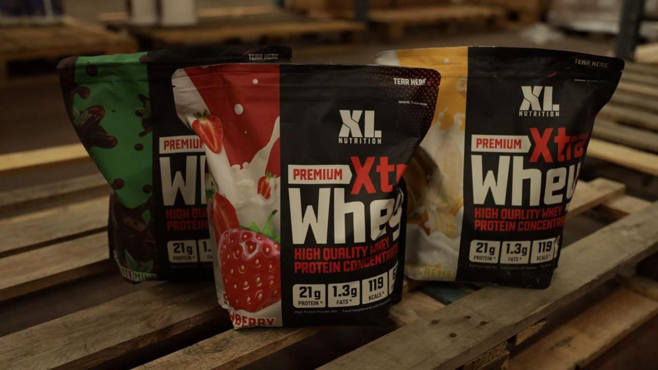 Three bags full of XL Nutrition Whey protein on a wooden pallet. On the black bags which are sealed at the top there is a photo of a strawberry, another has a banana and another is chocolate mint. It says Premium Xtra Whey on each bag.