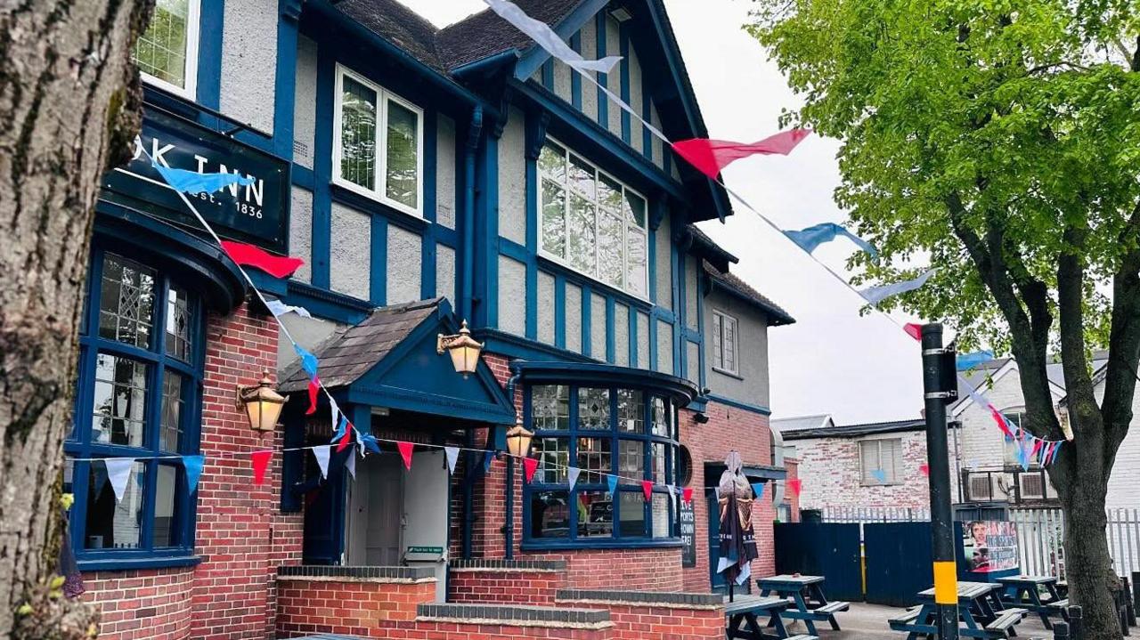 A black and grey pub has colourful bunting outside in the beer garden. Black benches are in the garden. 