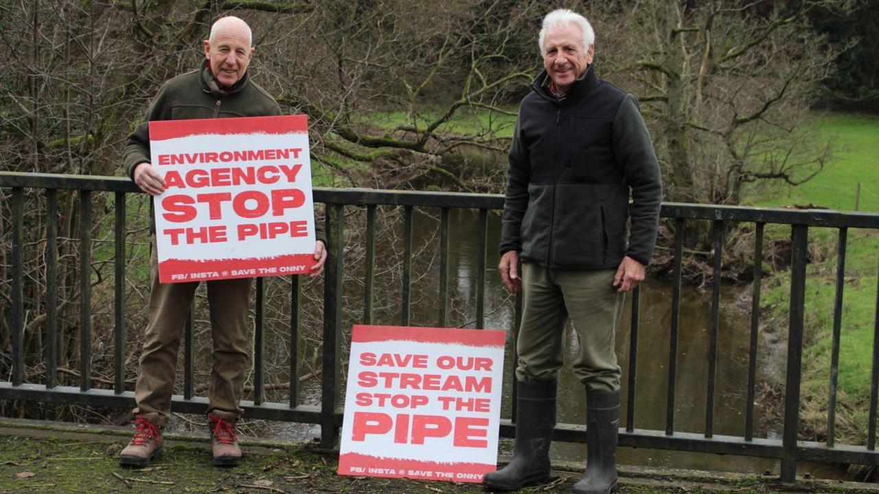 Two men stand on a bridge over a river with metal railings and a grass and tree-lined riverbank beyond. The men wear khaki trousers and dark green fleeces. The man on the left, who is bald, holds a sign that says "Environment Agency Stop the Pipe". A second sign is propped on the floor between the two men. The man on the right is grey-haired.