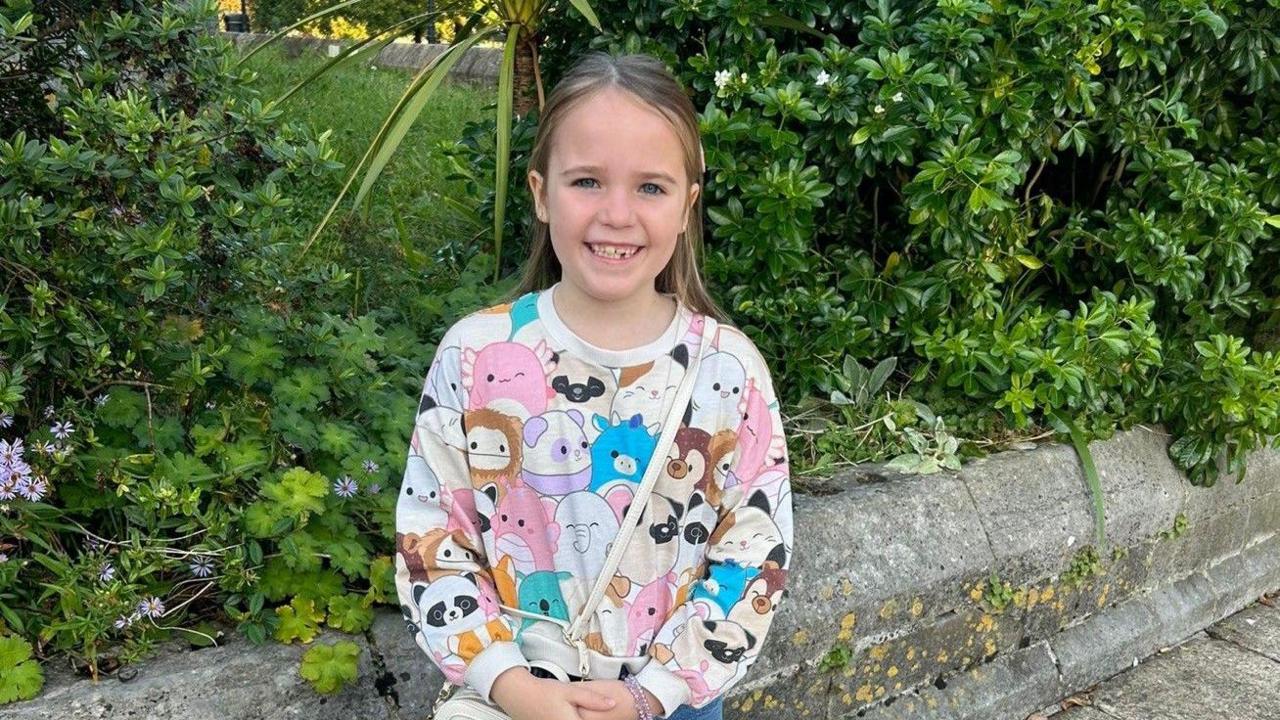 Seven-year-old Ellie stood surrounded by green bushes, she is wearing a long-sleeved top with different cartoon animals on, she is smiling.