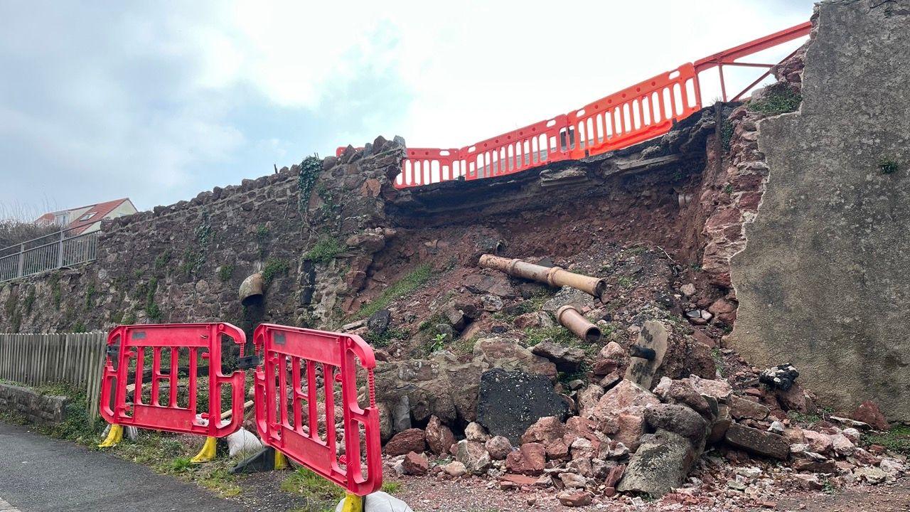 The landslip on Ringmore Road in Shaldon. A partially collapsed high stone wall has rubble and debris scattered at its base. Pipes and other structural elements have also been exposed. Bright red plastic safety barriers have been placed around the damaged area to prevent access and indicate danger.