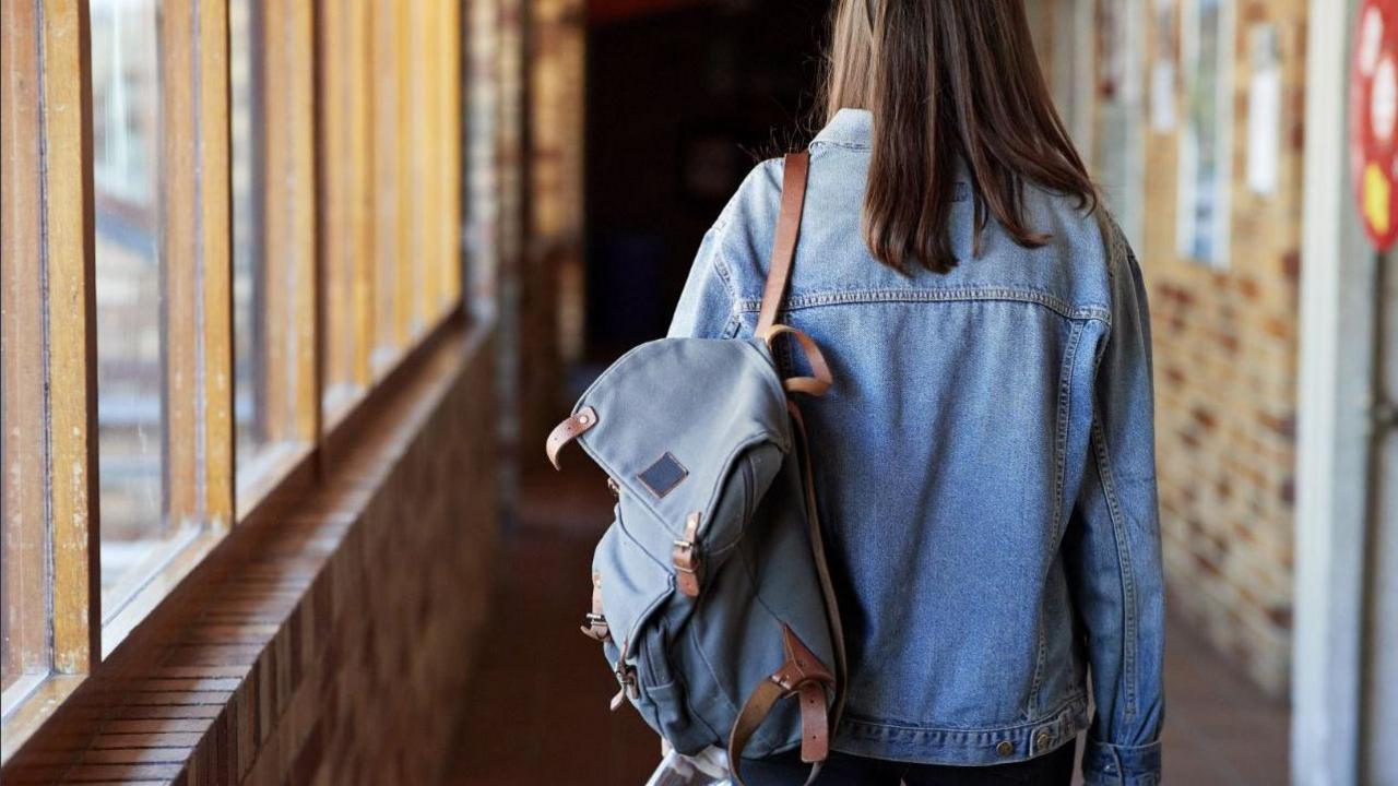 Rear view of young woman with backpack (rucksack or satchel drapped over shoulder) walking in corridor