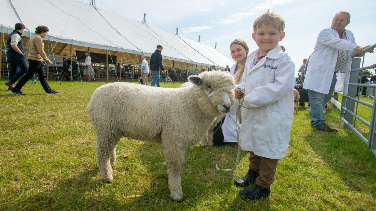 A young boy with short blond hair wearing a long white jacket and holding the lead of a sheep standing next to him on grass. A man and woman also in white jackets are smiling behind him and you can see a long marquee and people walking around on the left.