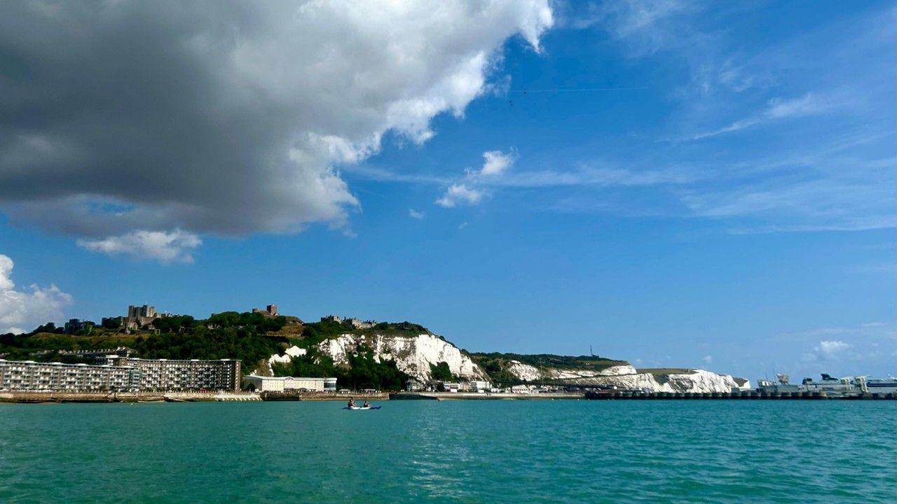 White cliffs on a seashore meet turquoise water under a dark grey cloud with clear blue sky surrounding it.