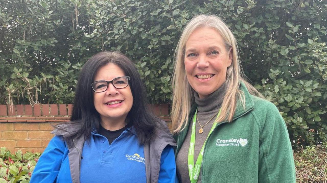 Anita Frith with long dark hair and glasses wearing a blue Cynthia Spencer top, and Jenine Rees with long blonde hair wearing a green lanyard and green Cransley Hospice top. They are standing in front of a hedge with a brick wall behind.