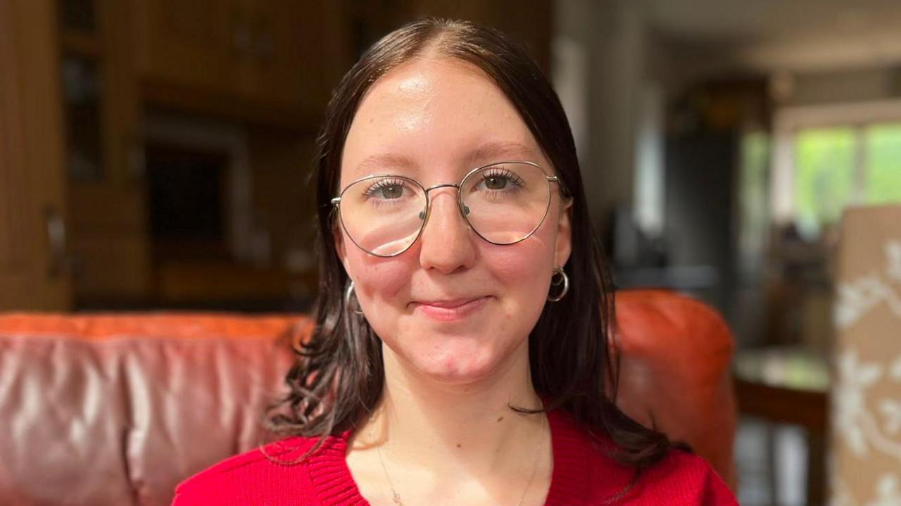 Lucy Eggink, a 20 year old white woman sitting on a brown leather chair. She has medium length brown hair and wearing a bright red cardigan and metal-framed glasses. 