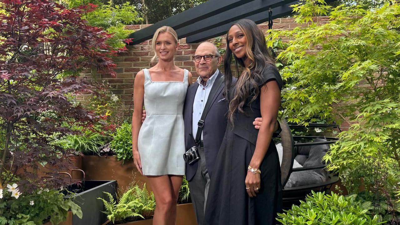Two women in dresses are stood in the garden, an older man wearing a shirt and blazer is stood between the with his arms around theirs waists