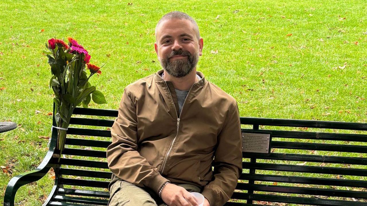 Geraint sitting on his wife's memorial bench  He is in a a brown collarless jacket with a zip and khaki trousers. There is a bunch of flowers tied to the bench.