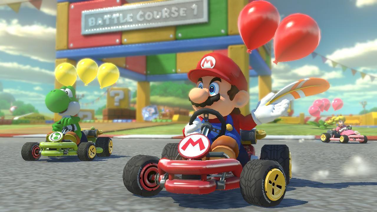 A screenshot from Mario Kart 8 Deluxe's Battle Mode shows Mario in a red kart and Yoshi in a green kart. They each have three balloons attached to their karts, and Mario holds a white and orange jump feather in his hand. The concrete track and grass areas of the arena stretch out behind them as Princess Peach races across the background.