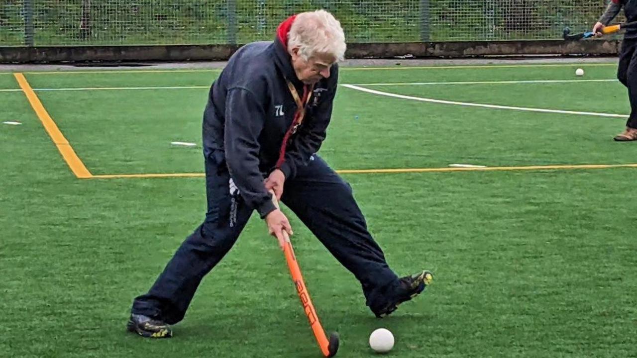 A man in a navy tracksuit playing hockey on astroturf