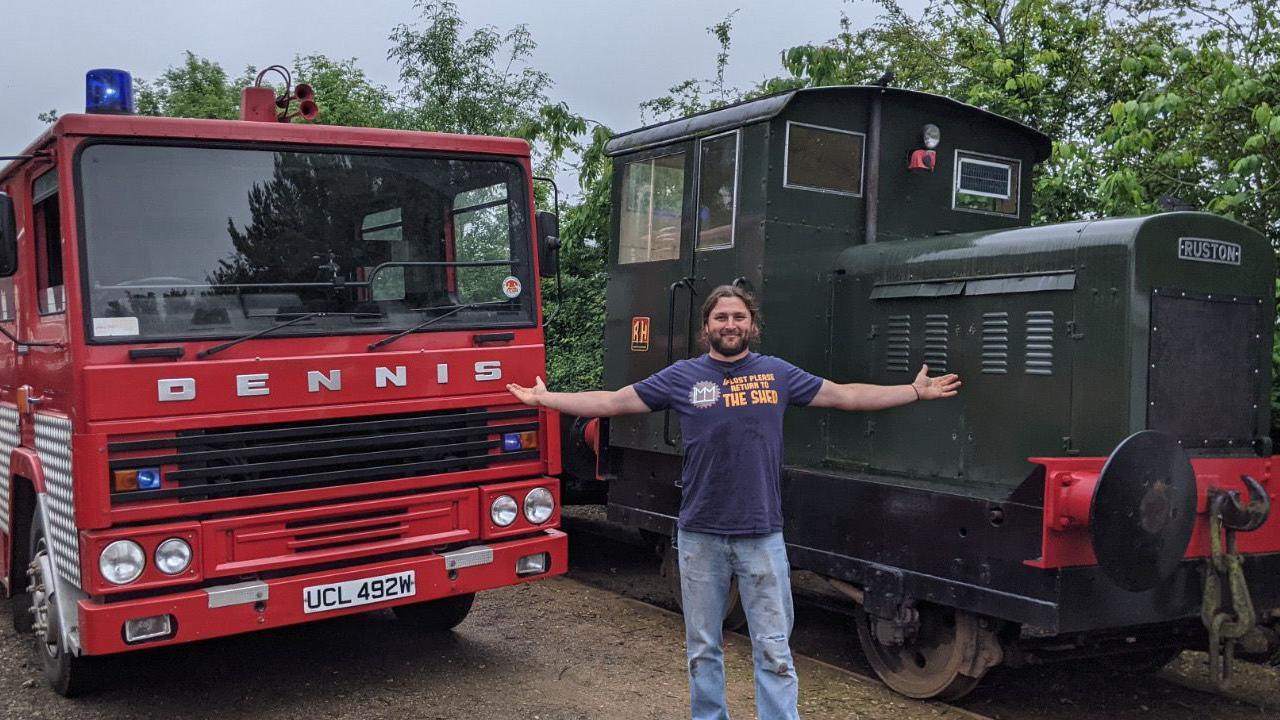 Lawrie Rose is pictured standing inbetween a green locomotive and red fire engine parked next to each other. He has long dark hair that is tied up and a dark beard. He is wearing a blue t-shirt with blue jeans. He has spread his arms out wide either side of him toward the vehicles. 