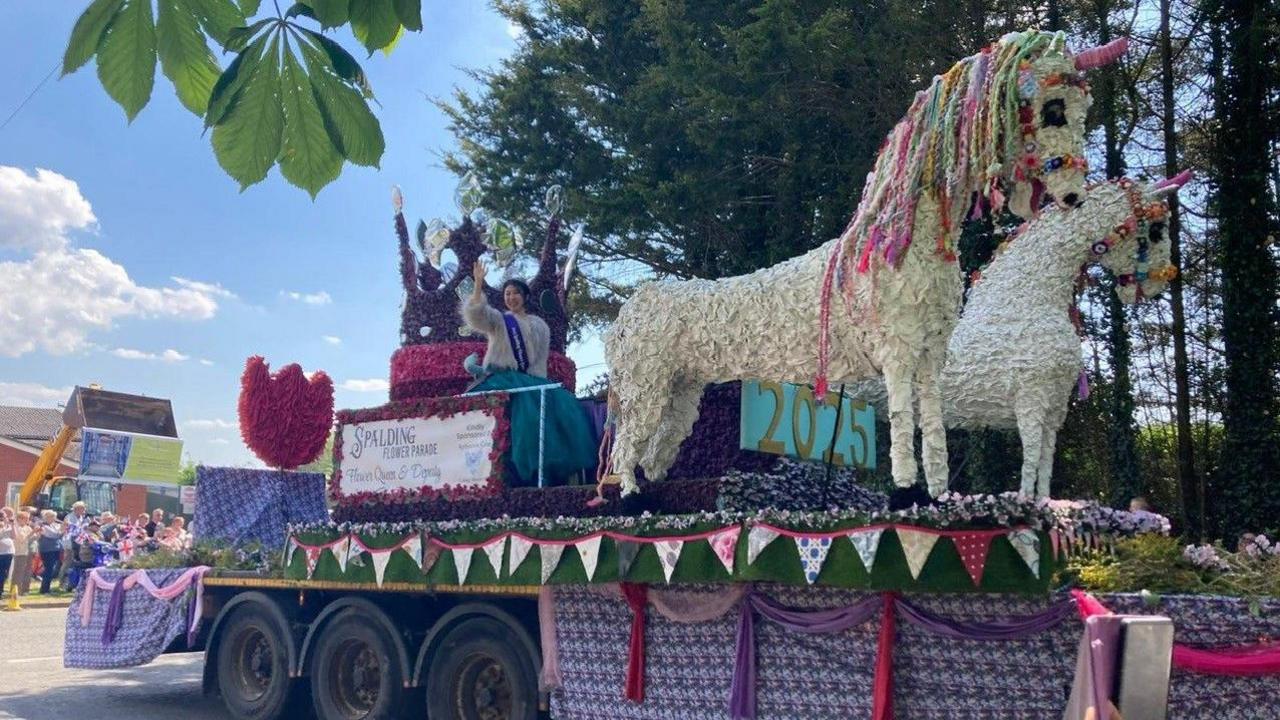 A large float on the flat bed of a lorry is adorned and draped with flags and flowers, including two large white horses that appear to pull a carriage which the flower queen sits in.