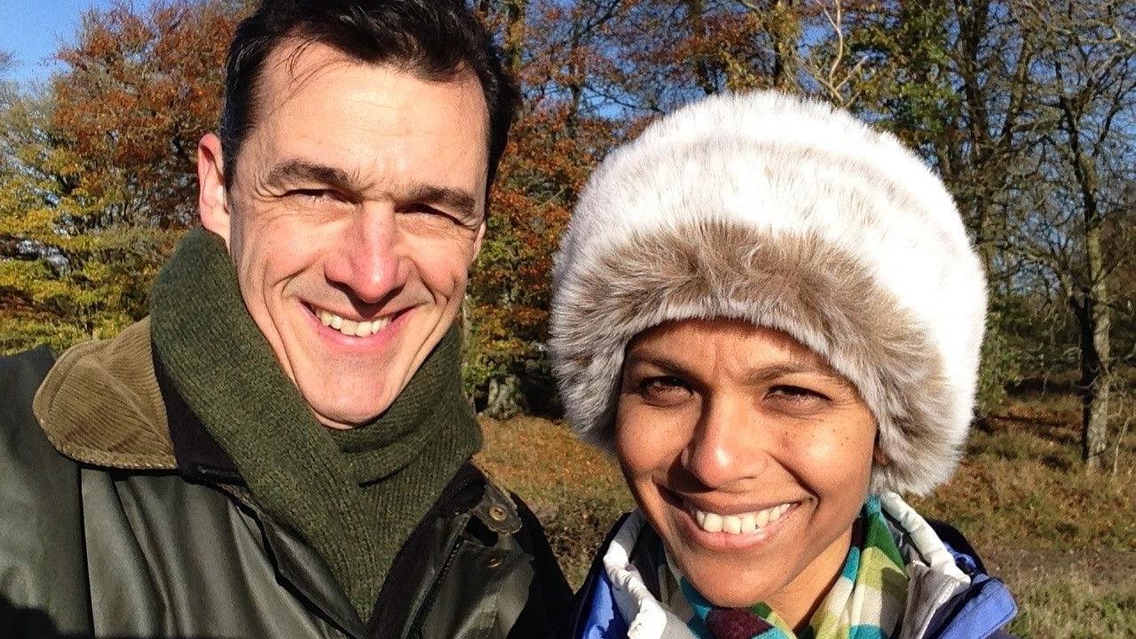 James and Alka Hughes-Hallett smile at the camera from outdoors, with trees behind them. It appears to be a cold day as They are in winter coats with scarves and Alka wears a fluffy hat